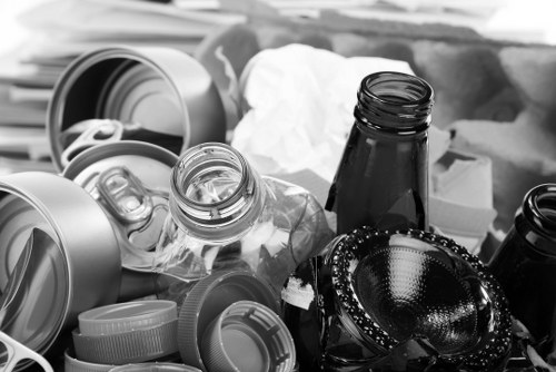 A collection of various discarded materials including a crumpled white tissue, several metallic and plastic bottles with their caps removed, and empty tin cans, some of which are open. The plastic bottles are transparent with ridged surfaces, while the metal cans are smooth and shiny with labels removed, positioned on a surface that appears to be an outdoor or semi-outdoor area, possibly a driveway or back alley in Hertford. In the background, there are additional objects such as a cardboard box and larger containers, with some items stacked or leaning against each other, indicating a waste collection or rubbish removal scene managed by Commercial Waste Hertford. The overall environment is informal, involving the collection of household or packaging waste meant for disposal or recycling as part of local rubbish removal services.