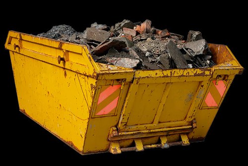 A large, rectangular yellow skip with metal reinforced edges, filled with a mixture of broken concrete, bricks, and rubble, situated outdoors on a paved surface. The skip has reflective pink safety markings on its rear panels and is positioned against a plain black background, indicating it is ready for waste collection or removal. The materials inside are dark grey and reddish-brown with rough textures, and the skip appears clean and well-maintained, consistent with industrial or commercial rubbish removal services in Hertford.