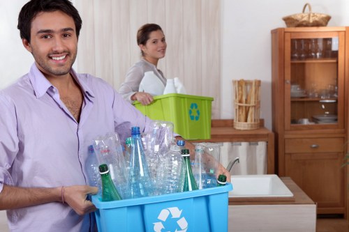 A young man holding a large blue recycling bin filled with empty plastic bottles, including clear and green-tinted bottles, in a domestic kitchen setting with wooden cabinets and a white countertop. Behind him, a woman is placing a smaller green recycling container on the counter. The kitchen has a neutral colour scheme with light wood cabinetry and a stainless steel sink, and the scene appears well-lit with natural or artificial light. The image illustrates rubbish disposal and recycling activity, relevant to commercial waste removal services in Hertford, showcasing proper recycling practices within a home environment, supported by Commercial Waste Hertford.