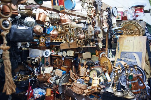A densely packed outdoor display of second-hand household items, including various brass and copper kitchenware such as teapots, trays, and utensils, along with a collection of ceramic vases, decorative plates, and small furniture pieces. The items are arranged on shelves, tables, and hanging from hooks, creating a cluttered yet organized scene typical of a vintage or thrift market. The materials show a mix of shiny metallic finishes and glazed ceramic surfaces, with some objects exhibiting intricate patterns and ornate designs. In the background, there are additional artifacts and items stacked and stored, suggesting a salvage or disposal setting near a commercial waste removal service in Hertford, accessible for disposal or recycling. The overall atmosphere reflects an outdoor market or warehouse environment focused on second-hand refurbishment or waste management, with natural daylight illuminating the scene.