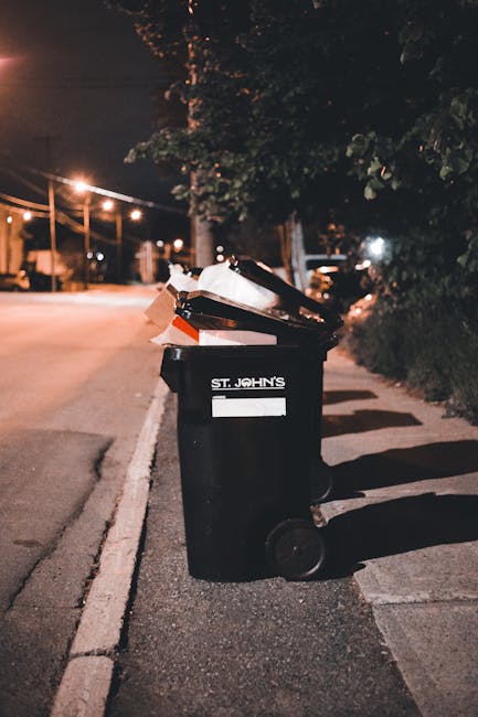 A black wheeled rubbish bin labeled 'ST. JOHN'S' positioned on a sidewalk beside a curb during nighttime. The bin is partially open and filled with various waste materials, including paper and plastic